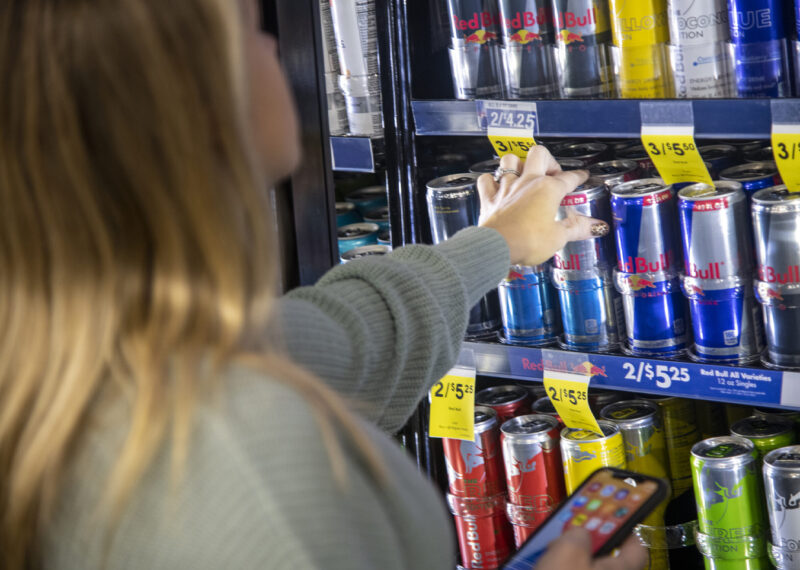 A woman picking up energy drink can and holding phone in other hand at CPG store