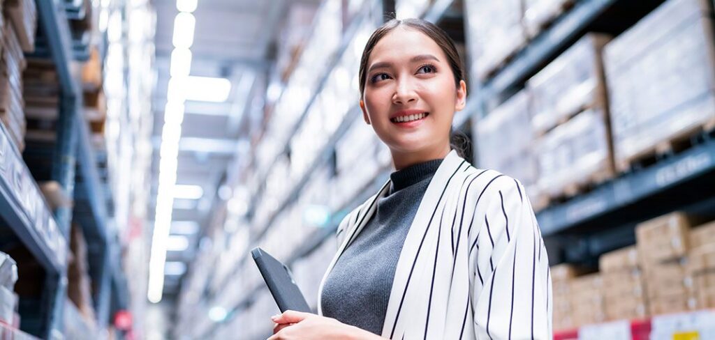 Businesswoman in back area of warehouse holding tablet and smiling.