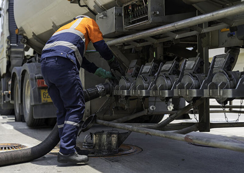 A fuel refinery worker is connecting a hose pipe to a fuel tanker