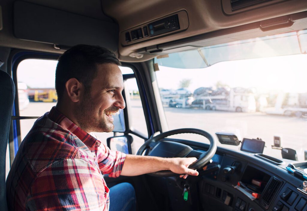 Longhaul trucker sitting in driver seat of truck cab smiling while looking out windshield.