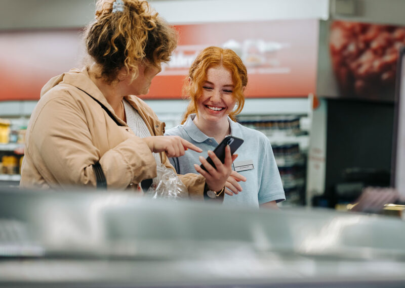 Smiling female employee at CPG store in front of POS while customer points to cell phone