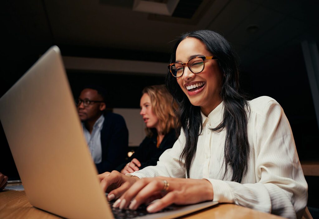 Close-up of woman with a big smile working on laptop in office with coworkers in background.