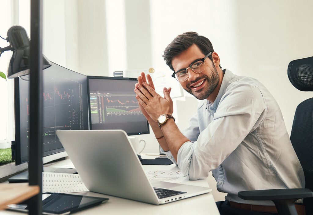 Happy bearded trader in formal wear and eyeglasses looking at camera and smiling while sitting in his modern office