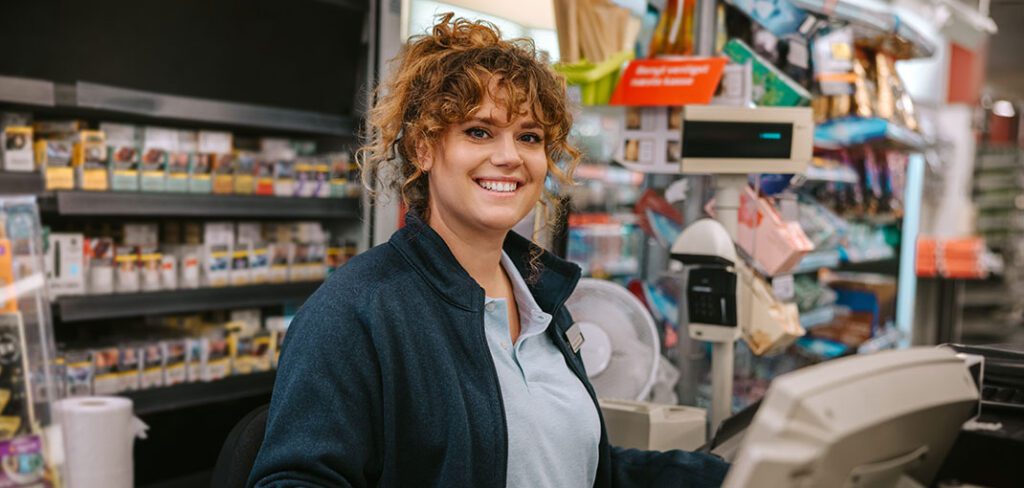 Smiling female cashier standing at checkout in front of retail POS system.