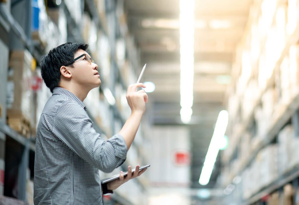 Male warehouse worker taking inventory of boxes on shelves using tablet and stylus.