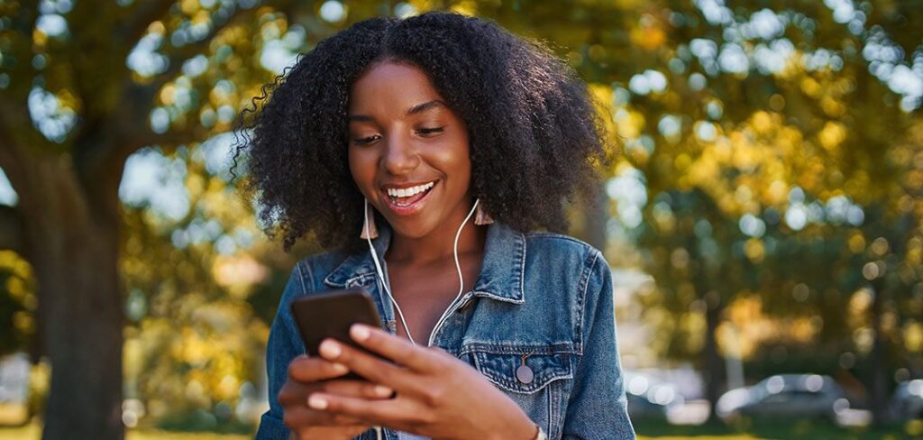 Portrait of smiling young african american woman listening to music on earphone using mobile phone in the park.