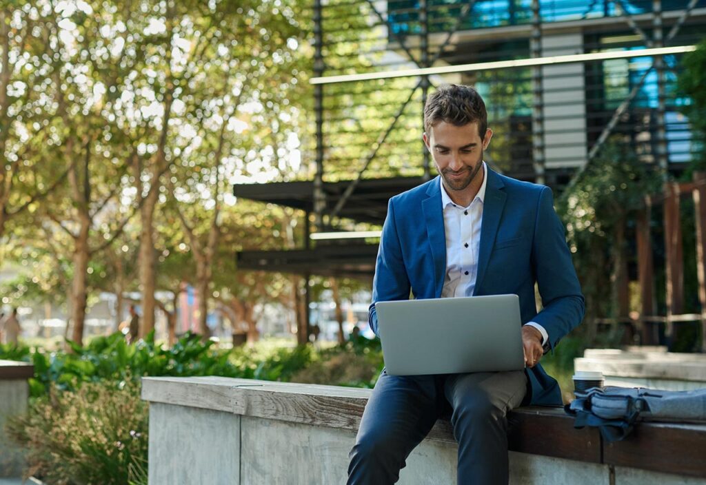 Business man in suit sitting in front of office building working on laptop.