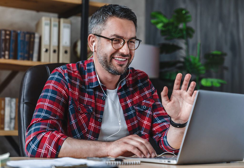 Man working on laptop from home waving to coworkers on team chat.