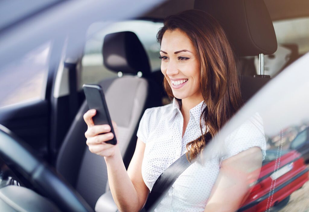 Woman holding cell phone smilling while in drivers seat of car.