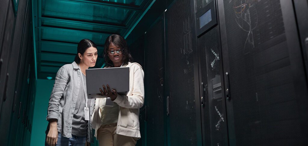 Waist up portrait of two female IT engineers setting up server network via laptop while working in data center.
