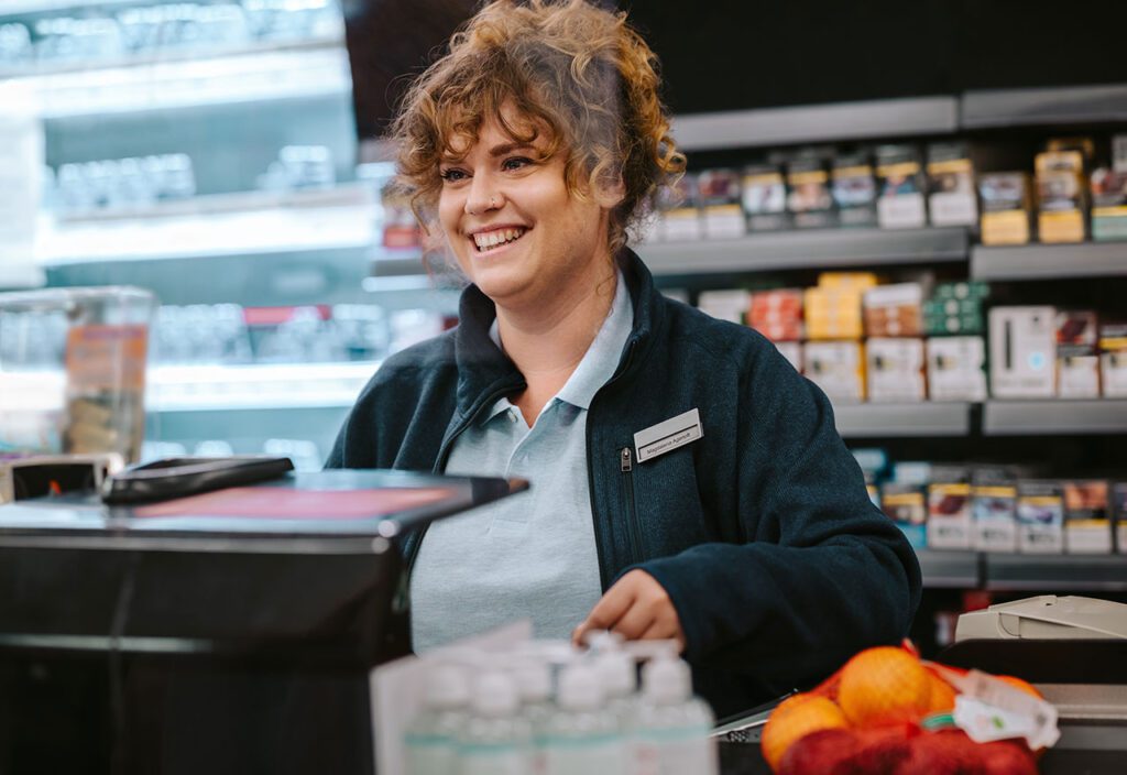 Cashier at conveinece retail location standing in front of consumer packaged goods smiling.