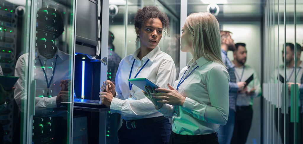 Female data center employees standing and talking in front of secure network servers.