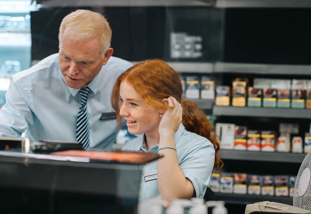 Manager training female cashier at CPG retail store.