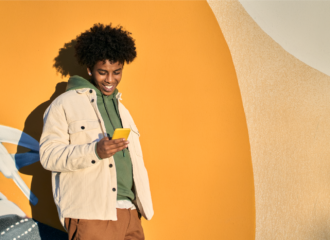 Happy smiling cool gen z young African American ethnic stylish hipster guy model standing at yellow city urban wall using cell phone mobile device, looking at smartphone, holding cellphone.