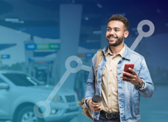 Smiling man holding coffee and mobile phone standing in front of background of convenience store forecourt with gasoline pumps
