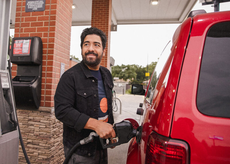 Man standing next to red SUV at convenience store, gas station filling up with fuel