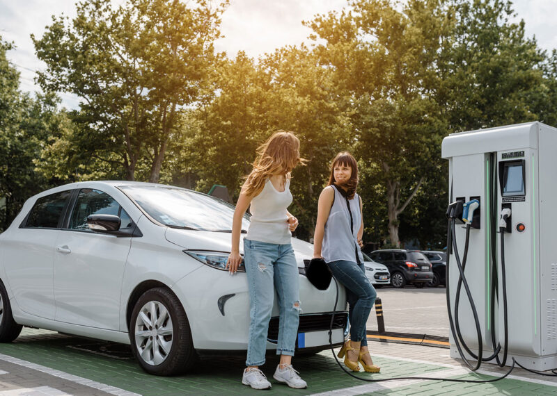 Two happy young beautiful women are talking to each other until their electric car is charging at the charging station situated in the car park.