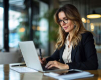 Busy mature mid aged business woman professional in her 40s writing notes finance report overview, lawyer checking document, working on laptop computer device sitting at desk in office