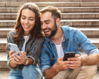 Two individuals sitting on outdoor steps, both engaged with their smartphones.