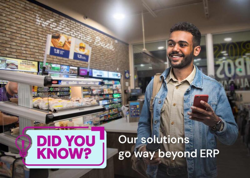 Smiling man holding a phone and a coffee cup inside a convenience store with a display of sandwiches in the background. Text overlay reads 'Did You Know? Our solutions go way beyond ERP.'