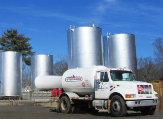 White tanker truck labeled Stringer Oil & LP Gas parked in front of three large vertical storage tanks under a clear blue sky.