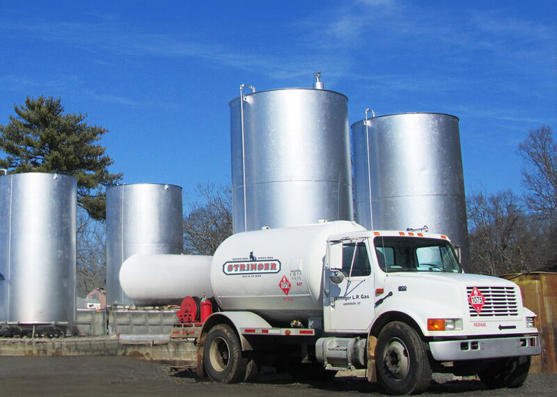 White tanker truck labeled Stringer Oil & LP Gas parked in front of three large vertical storage tanks under a clear blue sky.