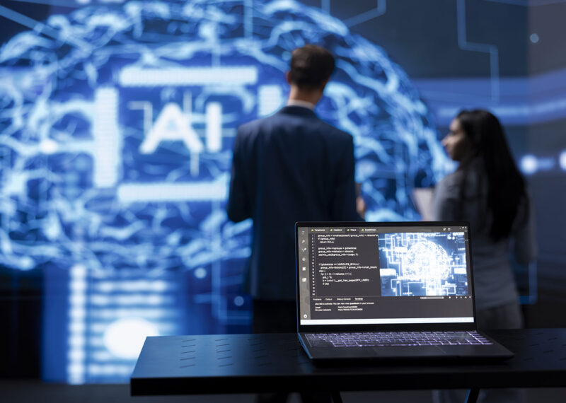 Laptop displaying AI code and brain visualization in a cybersecurity lab, with professionals discussing in the background, symbolizing AI-powered threat detection and proactive defense.