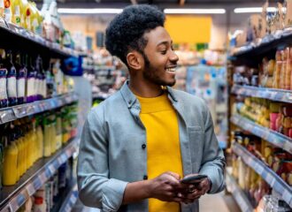 Smiling man shopping in a convenience store aisle, holding a mobile phone while looking at shelves stocked with product offers from Consumer Packaged Goods brands, illustrating convenience retail and c-store shopping experiences.