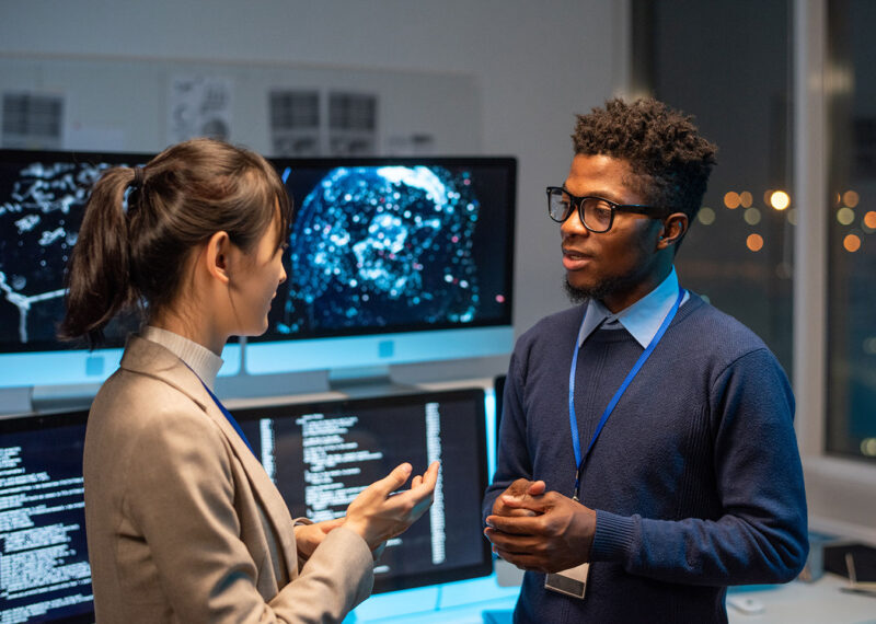 Two cybersecurity professionals discussing threat intelligence in a security operations center, with digital world maps and data visualizations on screens.