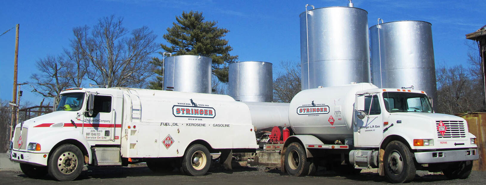 Stringer Oil Company fuel and propane delivery trucks parked in front of large silver fuel storage tanks under a clear blue sky.