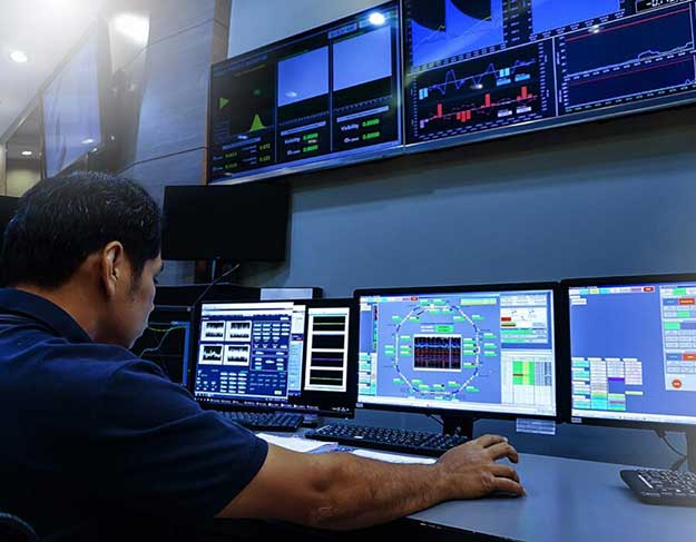 Person working at a cybersecurity monitoring center with multiple screens displaying real-time data, graphs, and system analytics.