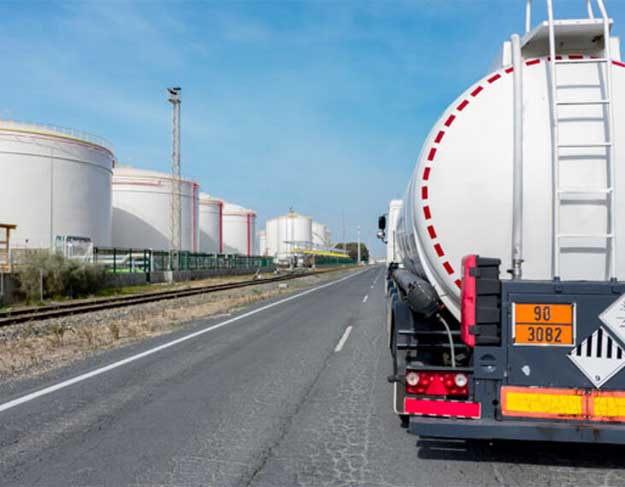 Fuel tanker truck driving on a road near large fuel storage tanks at an industrial facility under a clear blue sky.