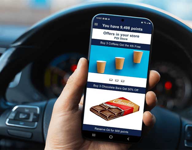 Person holding a smartphone inside a car, displaying a digital loyalty rewards app with store offers and discounts on coffee and chocolate bars.