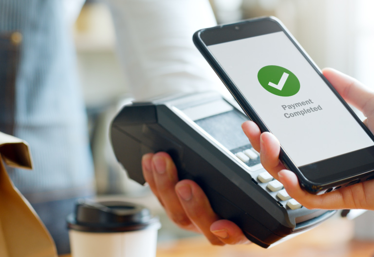Person completing a mobile payment using a smartphone at a point-of-sale terminal, representing PDI Digital Payment Solutions with secure in-app acceptance and integration to major payment processors and digital wallets.