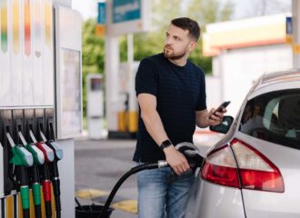 Bearded man refuelling car on gas station and looking into his smartphone. Man compares fuel prices