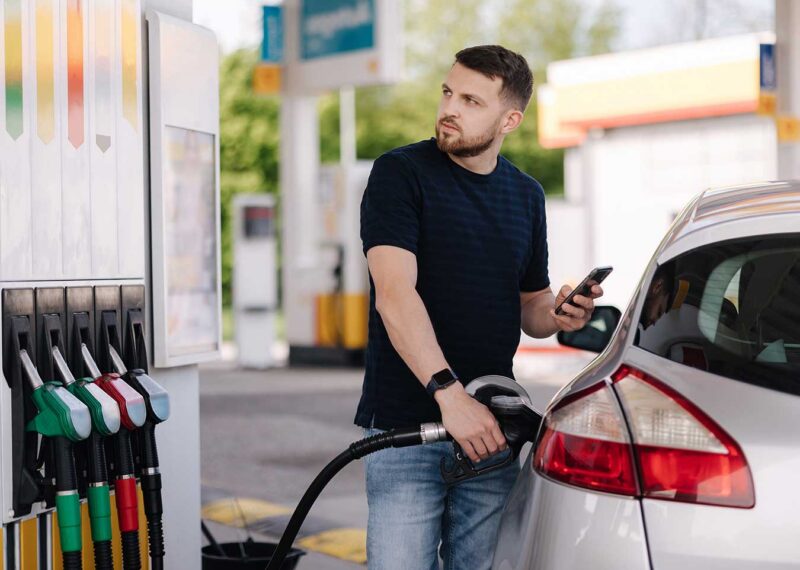 Bearded man refuelling car on gas station and looking into his smartphone. Man compares fuel prices