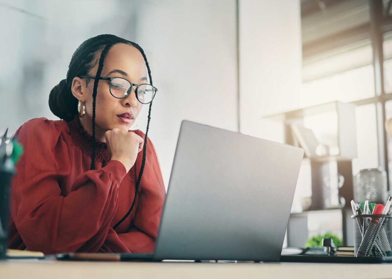 Woman in glasses sitting at a desk, looking thoughtfully at her laptop.