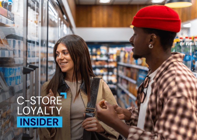 Two shoppers chat in front of a convenience store cooler aisle, with a 